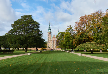Rosenborg castle long This landscape photograph features Rosenborg Castle, a notable example of architecture in Copenhagen, Denmark. Captured in the late morning during autumn, the image shows the castle set against a backdrop of partly cloudy skies and surrounded by the vibrant colors of seasonal foliage. The well-maintained lawns and tree-lined pathways demonstrate the orderly design typical of historic Copenhagen parks. Rosenborg Castle, with its distinctive towers and ornate brickwork, stands prominently in the center, making it a key landmark in the image.